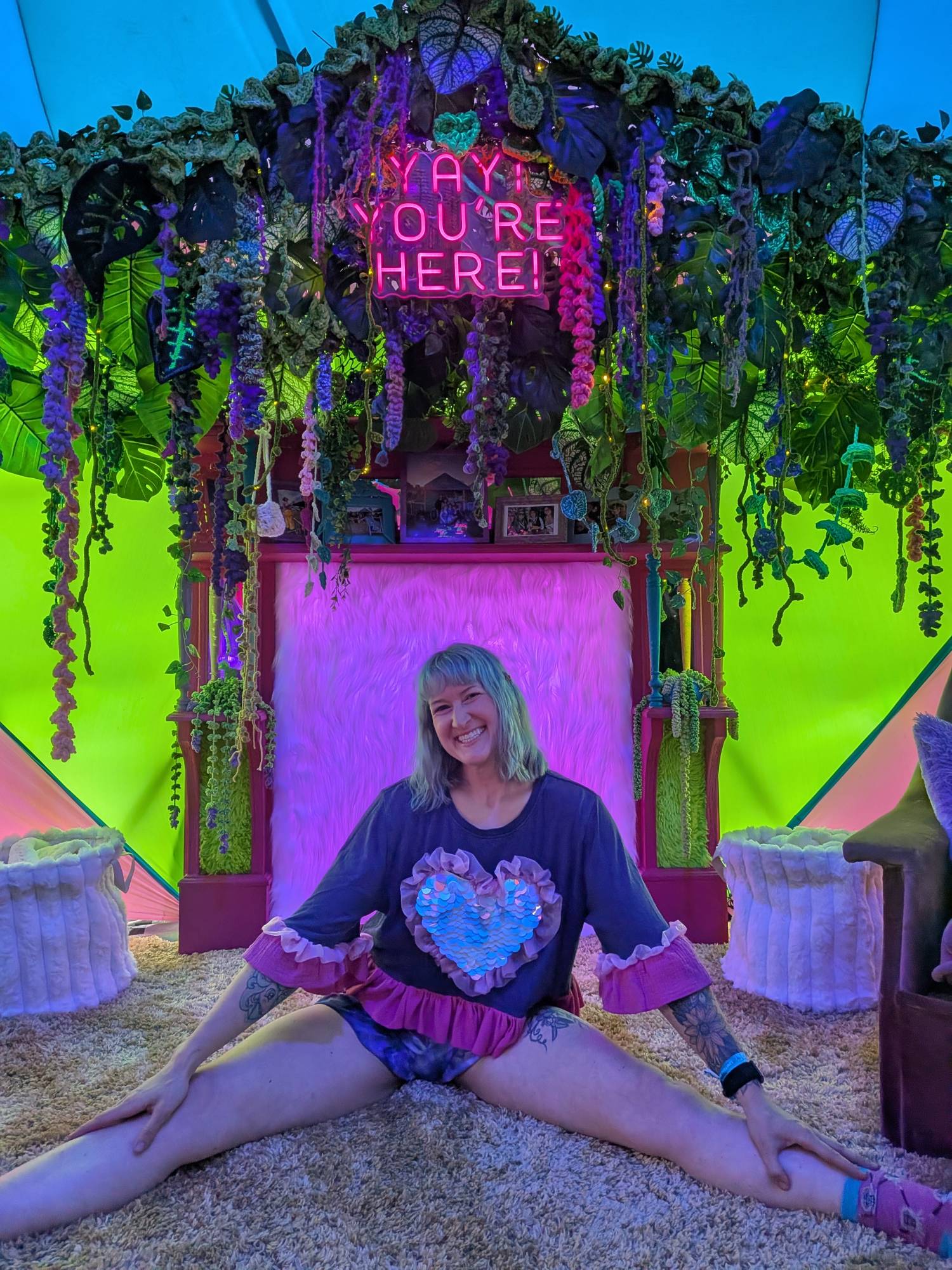 Jess doing the splits in front of the touch-reactive LED fireplace, draped in a crochet plant wall under a neon ‘Yay! You’re here!’ sign