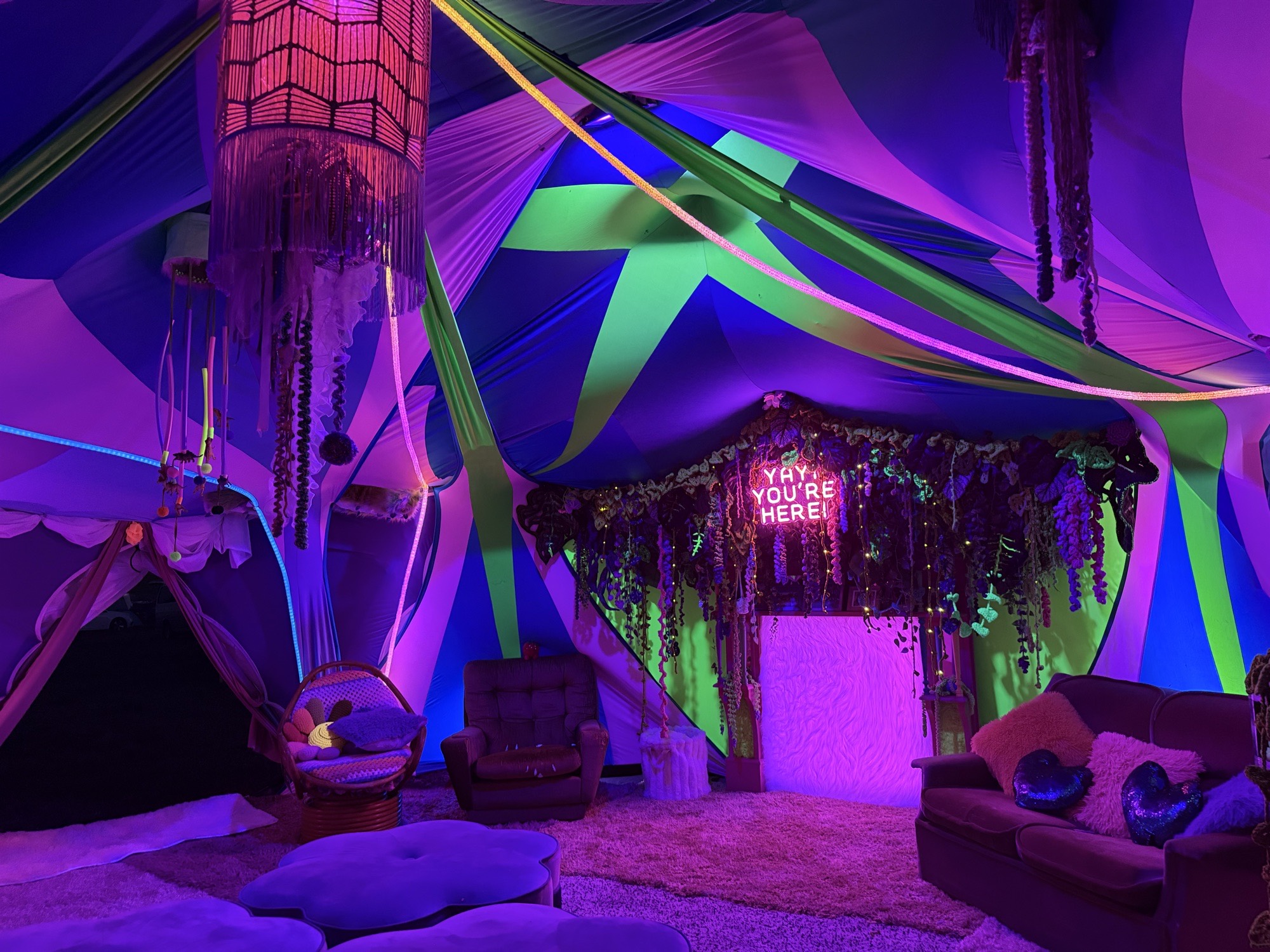 The lounge corner of the Aunty Maria’s tent glowing purple, with a couch, egg chair and the fireplace under its neon sign