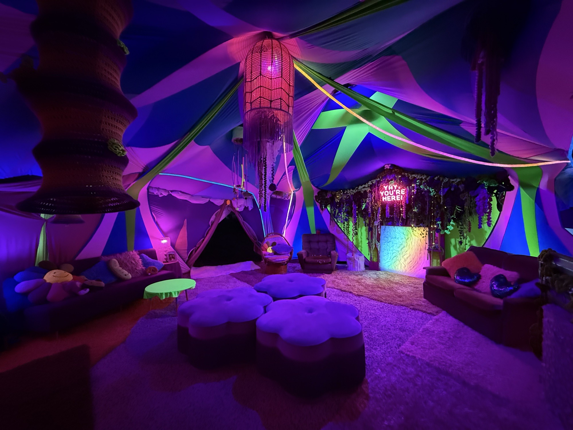 Inside the Aunty Maria’s tent with the lights down: purple and green draped ceiling, glowing lanterns, flower-shaped ottomans and the neon-lit fireplace at the back