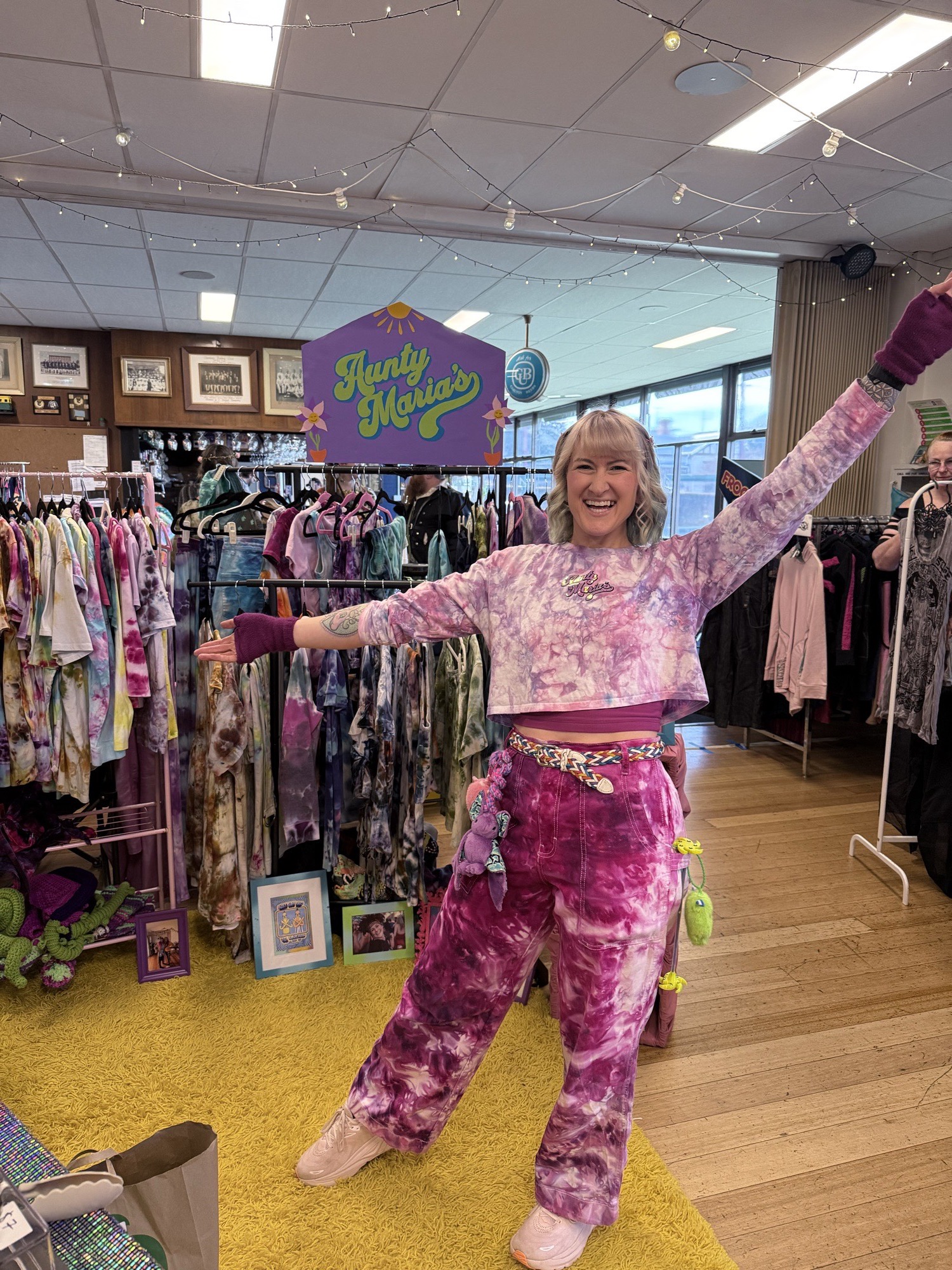 Jess in tie-dye with arms outstretched at the Aunty Maria’s market stall, racks of colourful clothes behind her