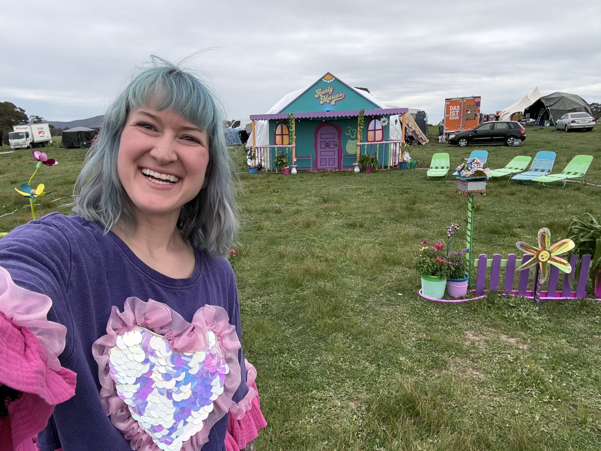 Jess grinning in a selfie with the finished Aunty Maria’s house behind her in a grassy campground