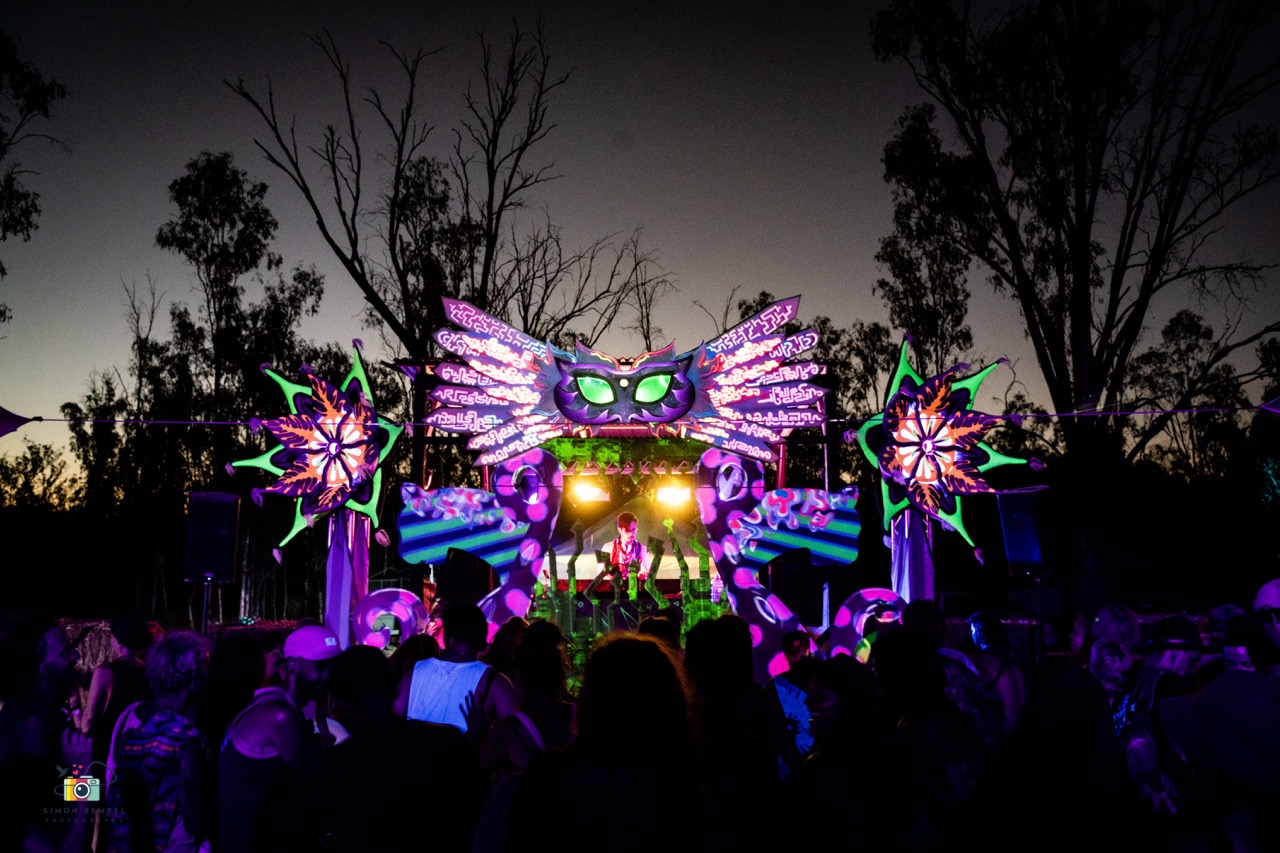 A UV-painted stage at dusk: a winged mask with glowing green eyes over the DJ booth, flanked by two spiky flower sculptures, crowd in silhouette