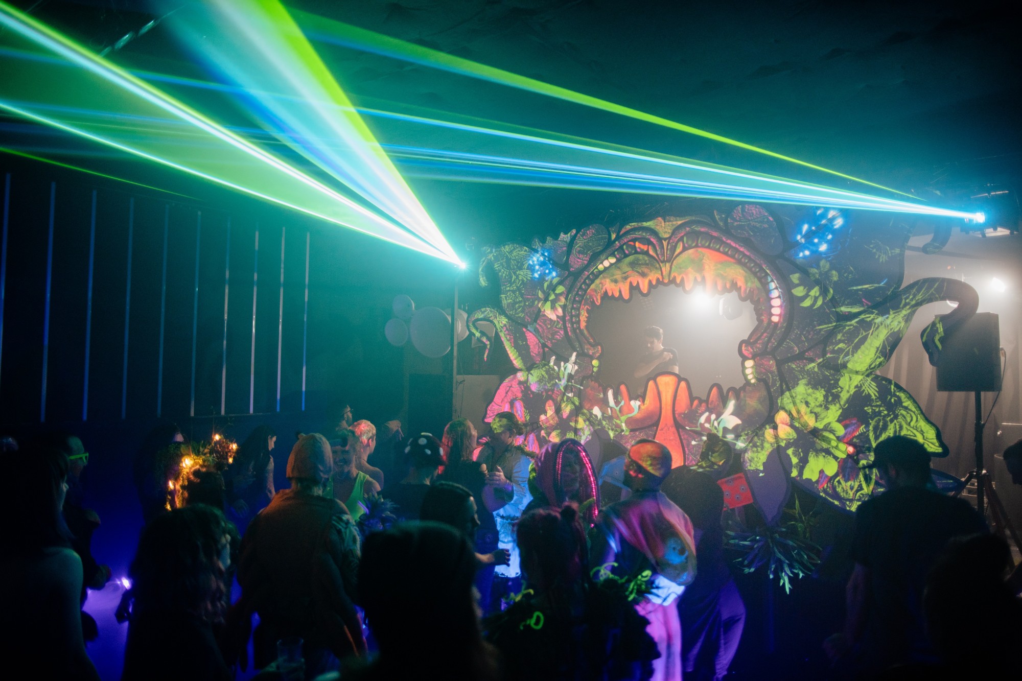 A crowd dancing under green lasers in front of a giant carnivorous plant mouth framing the DJ, lit with projection mapping