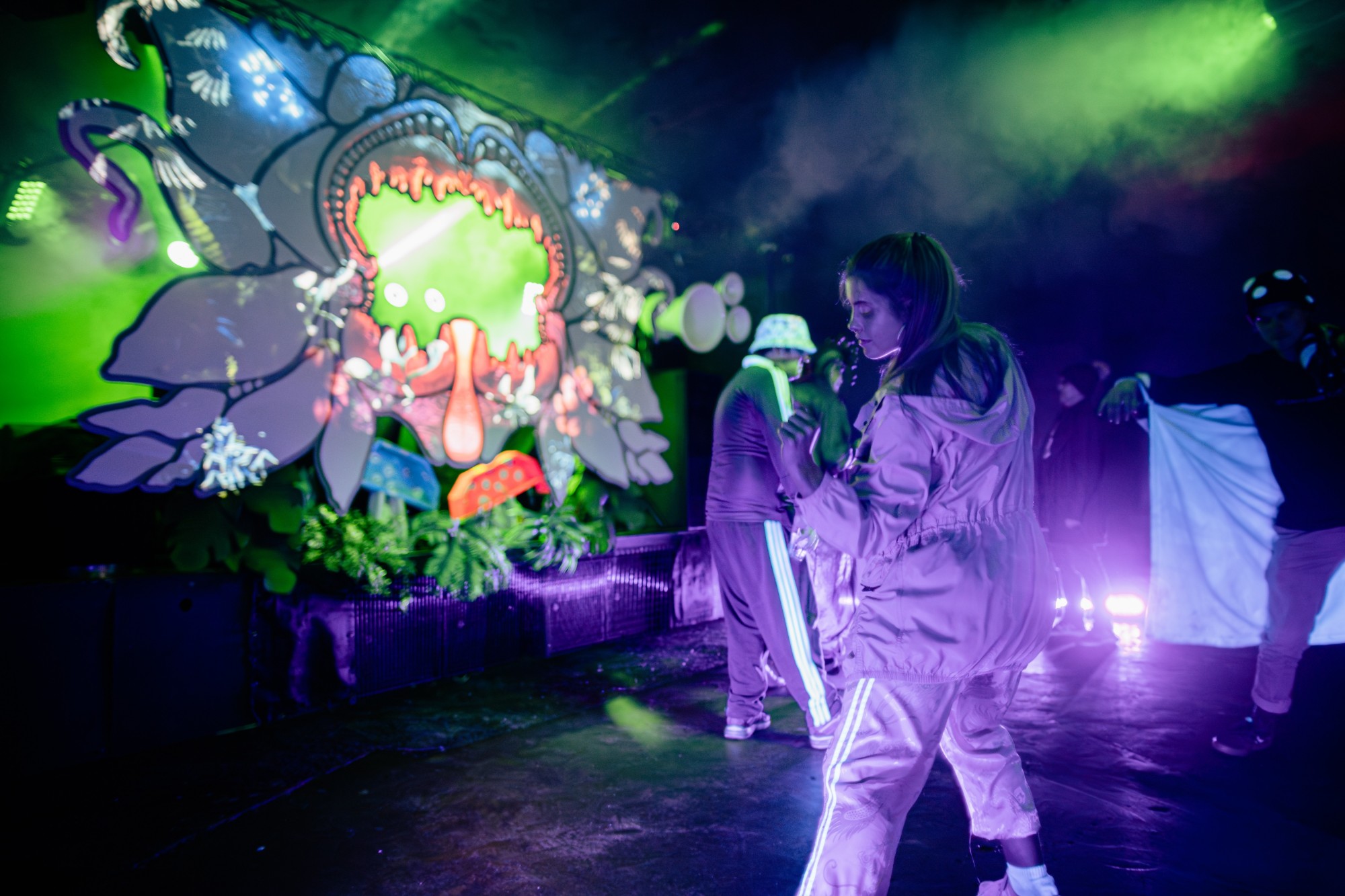 A dancer in a pale tracksuit lit purple, the carnivorous plant stage glowing green behind her