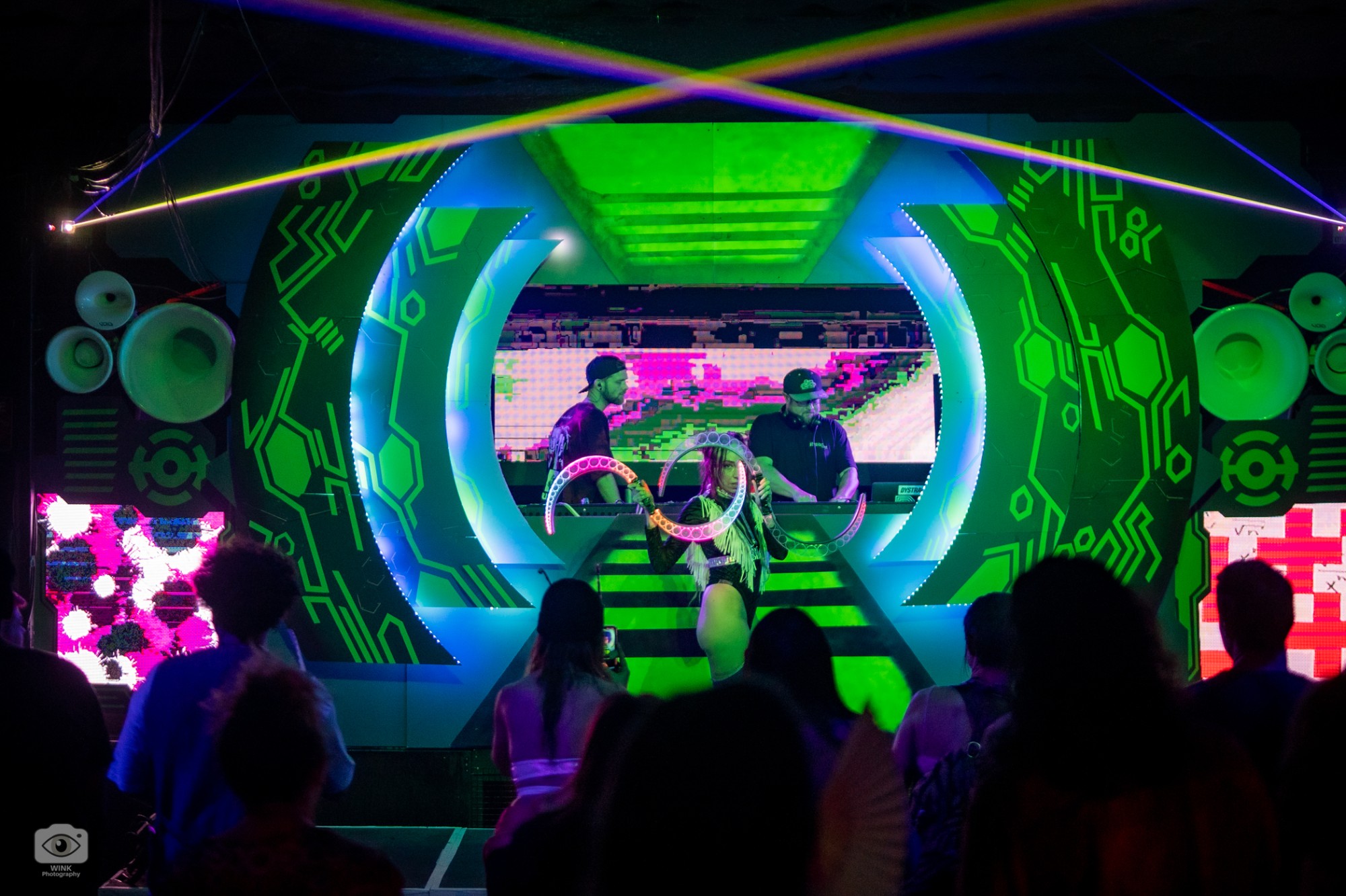 A circular portal stage with green circuit patterns and LED edging framing two DJs, lasers crossing overhead, a hoop performer out front