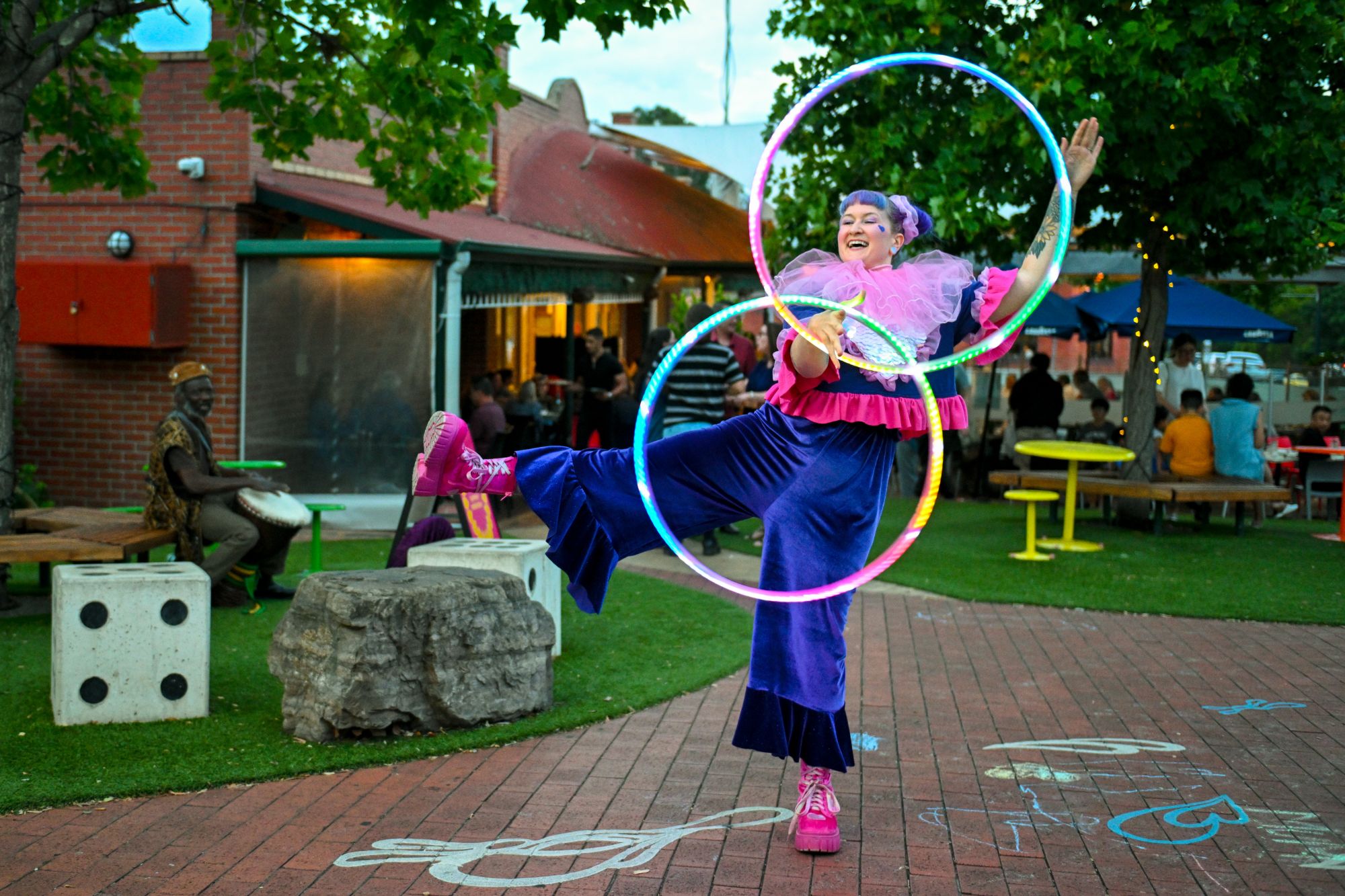 Jess in a purple velvet and pink ruffle costume kicking a leg high with two rainbow LED hoops in a courtyard, a drummer playing behind