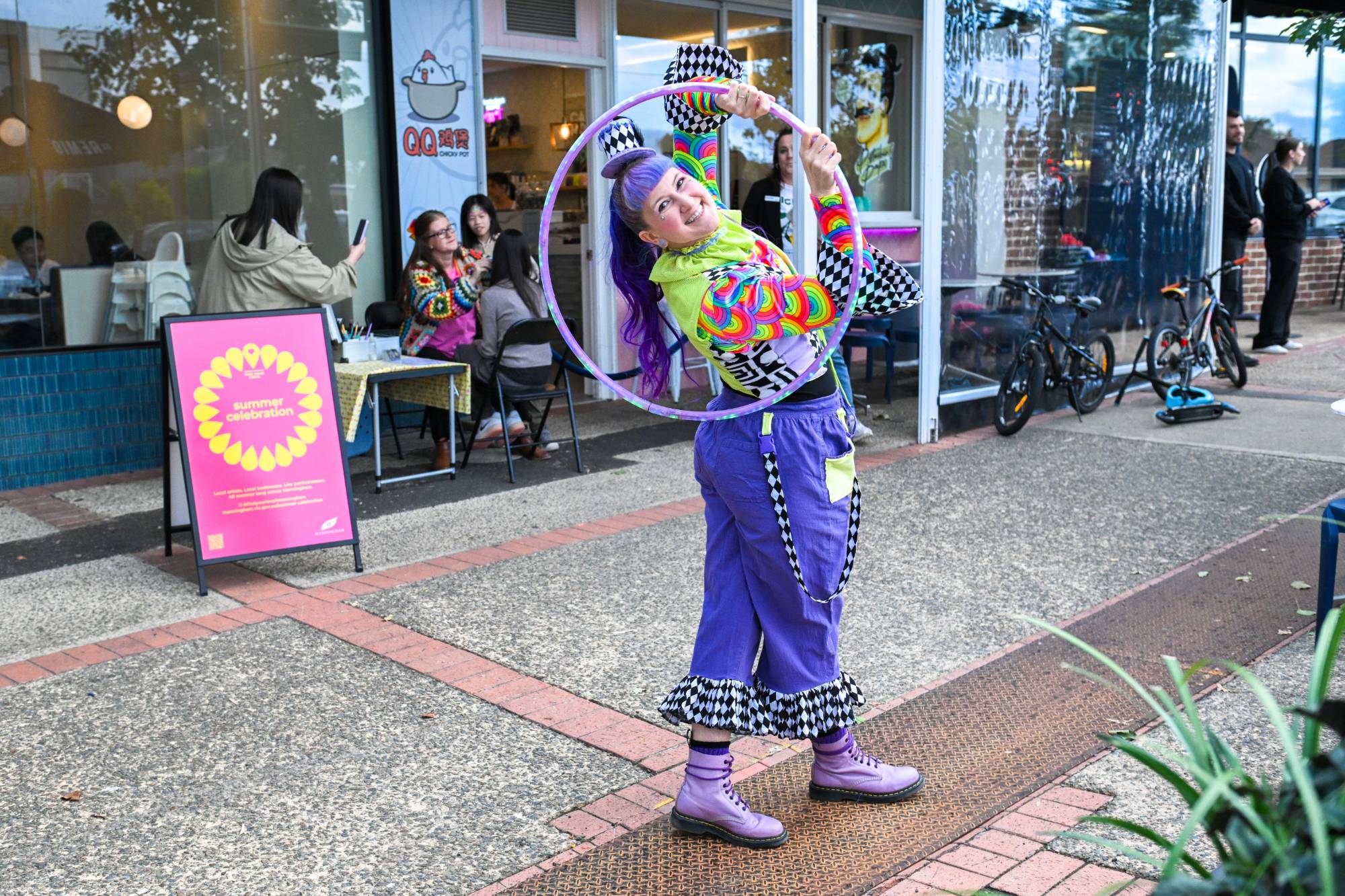 Jess in a rainbow and harlequin costume leaning back through a hoop on a footpath outside a cafe