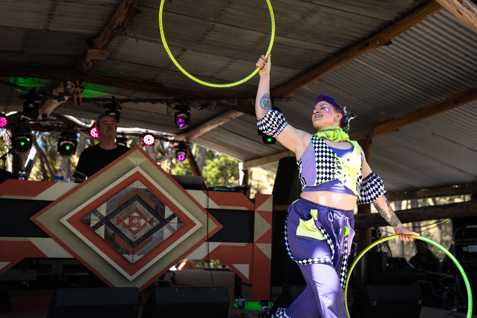 Jess in a purple and harlequin costume swinging two green hoops on a daytime festival stage
