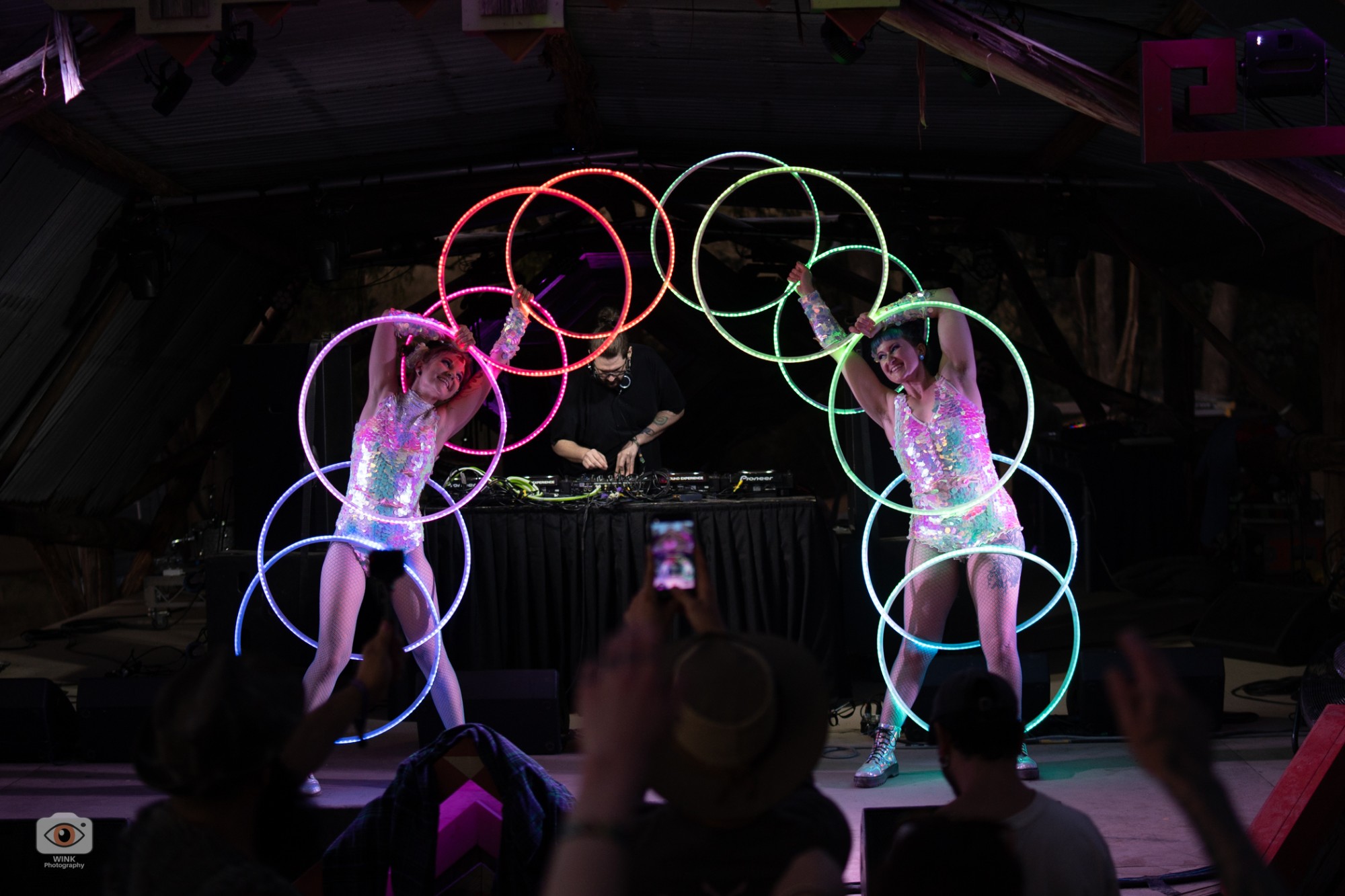 Two hoopers in matching sequinned bodysuits joining LED hoops into a rainbow arch over the DJ booth