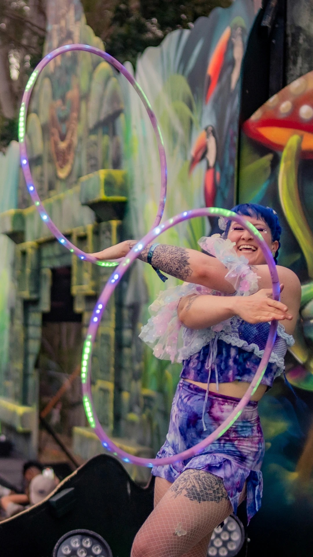Jess laughing while twinning two LED hoops in front of a jungle mural at a festival stage