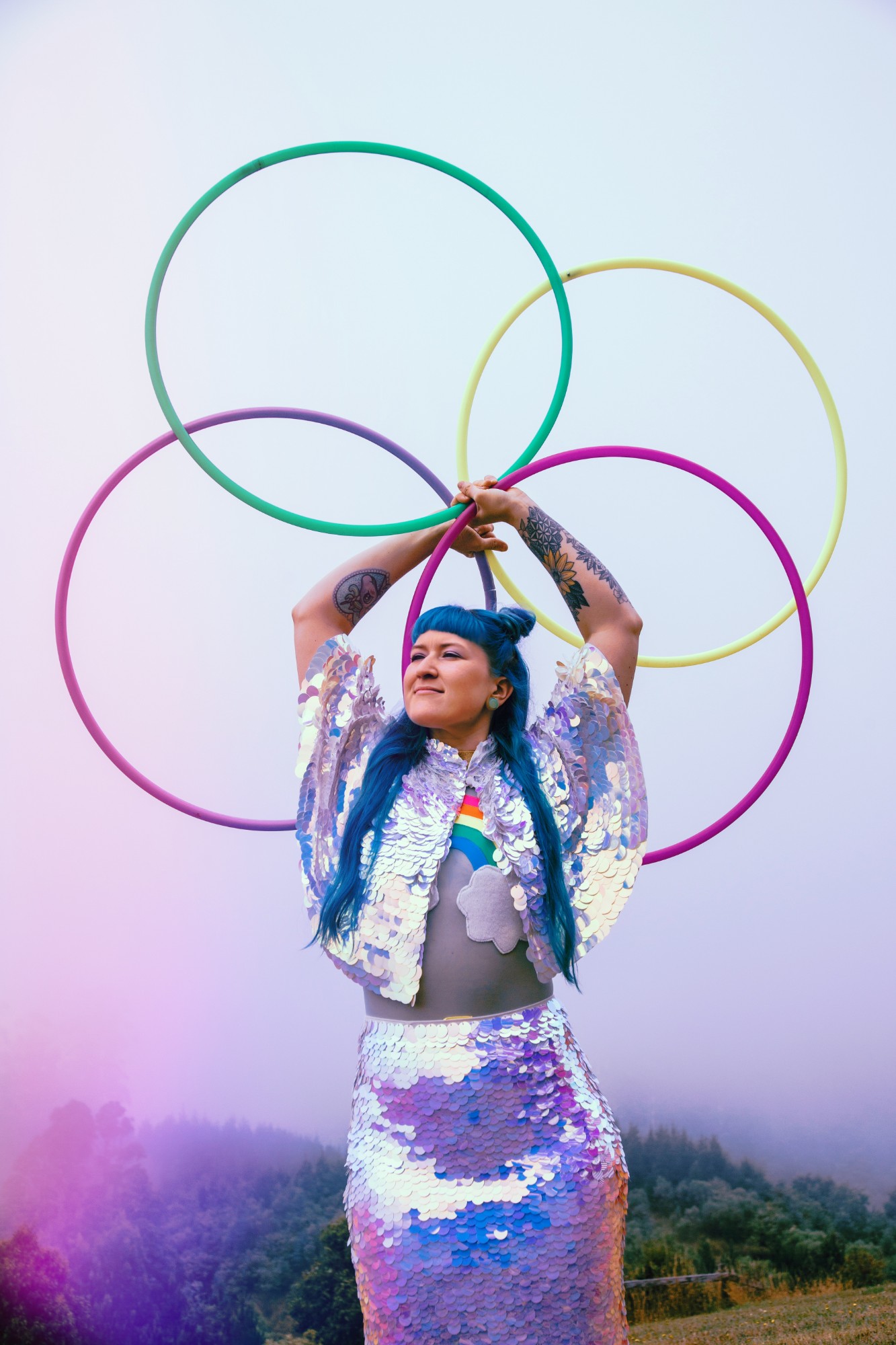 Jess in a sequinned outfit holding four pastel hoops overhead in a petal shape against a misty hillside