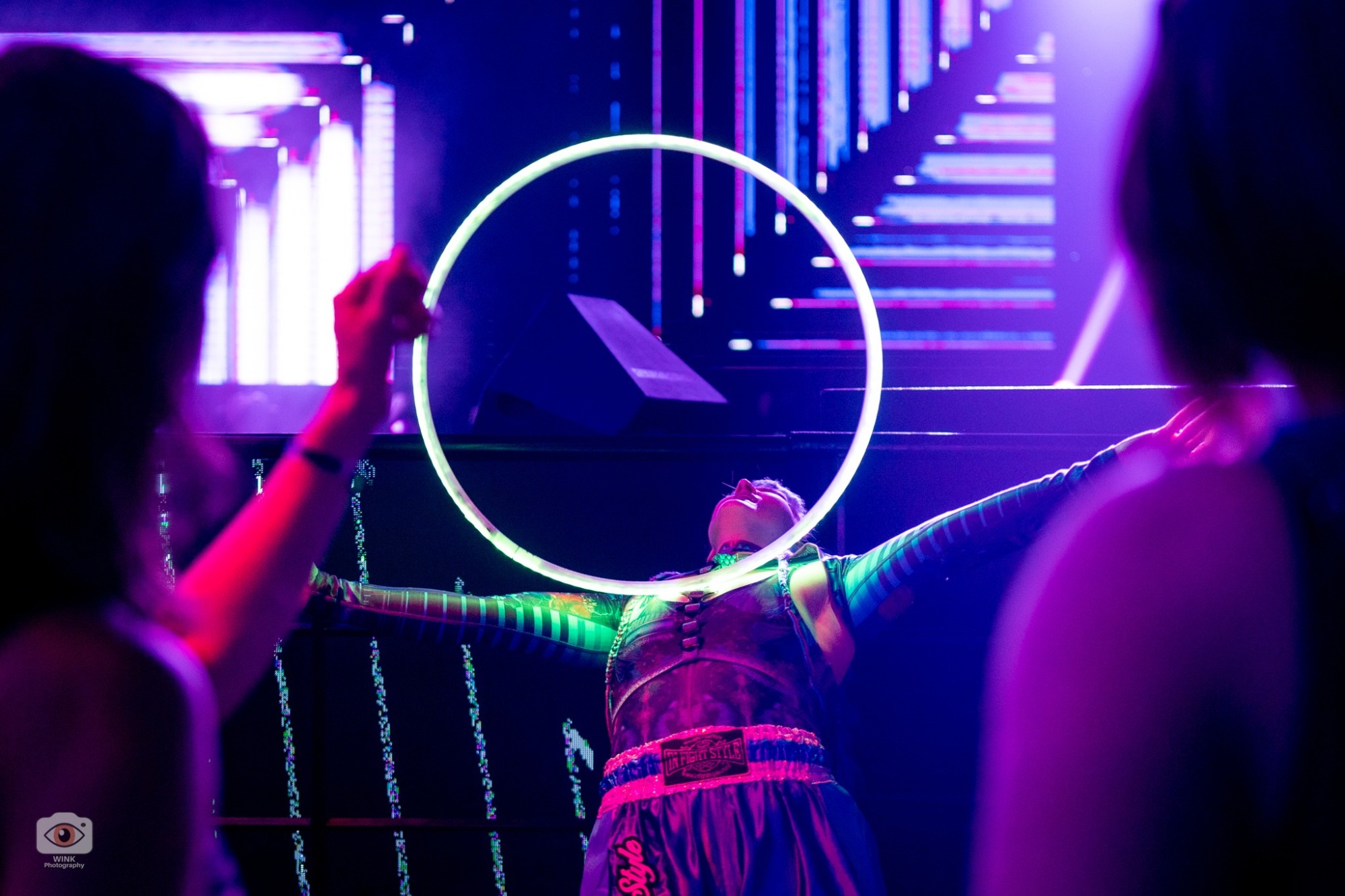 Jess leaning back with arms spread as a glowing hoop rises off her neck, LED screens behind