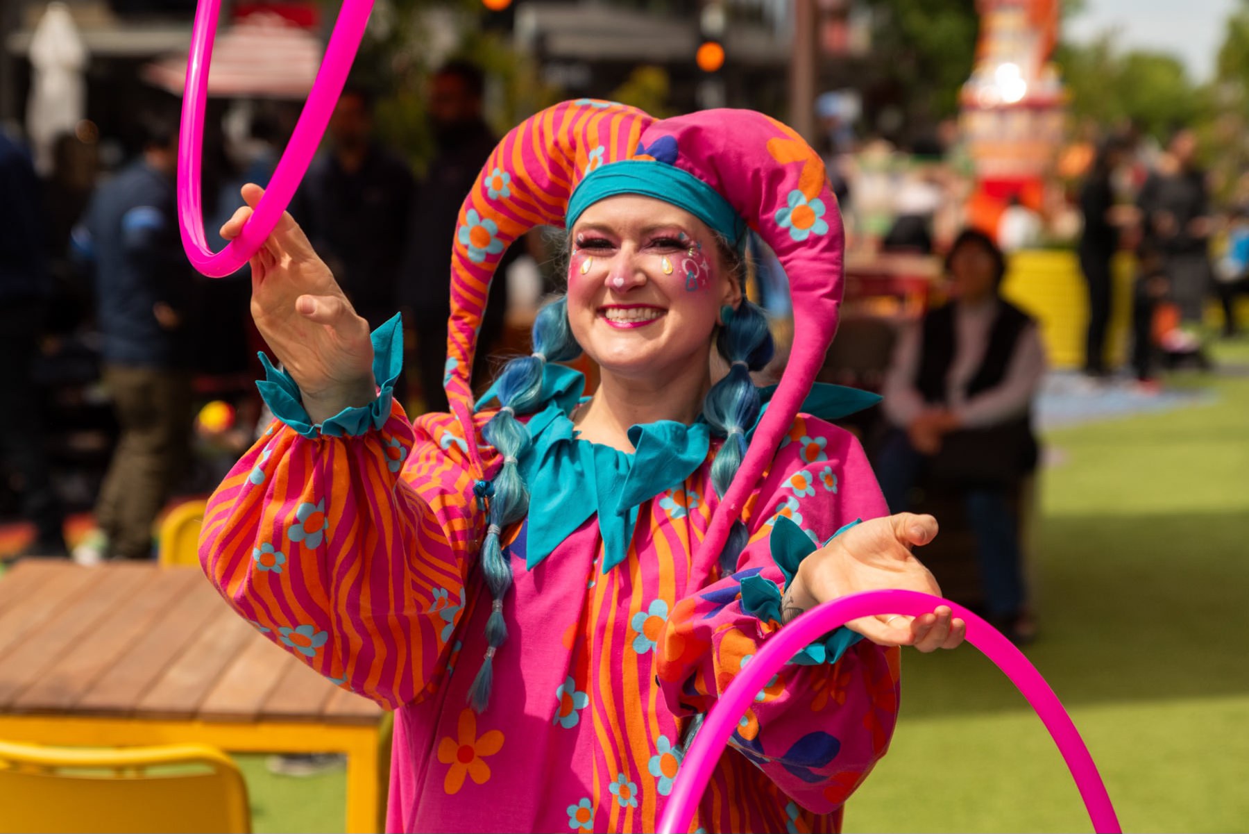 Jess smiling in a pink and orange jester costume with blue braids, tossing a pink hoop at a daytime community event