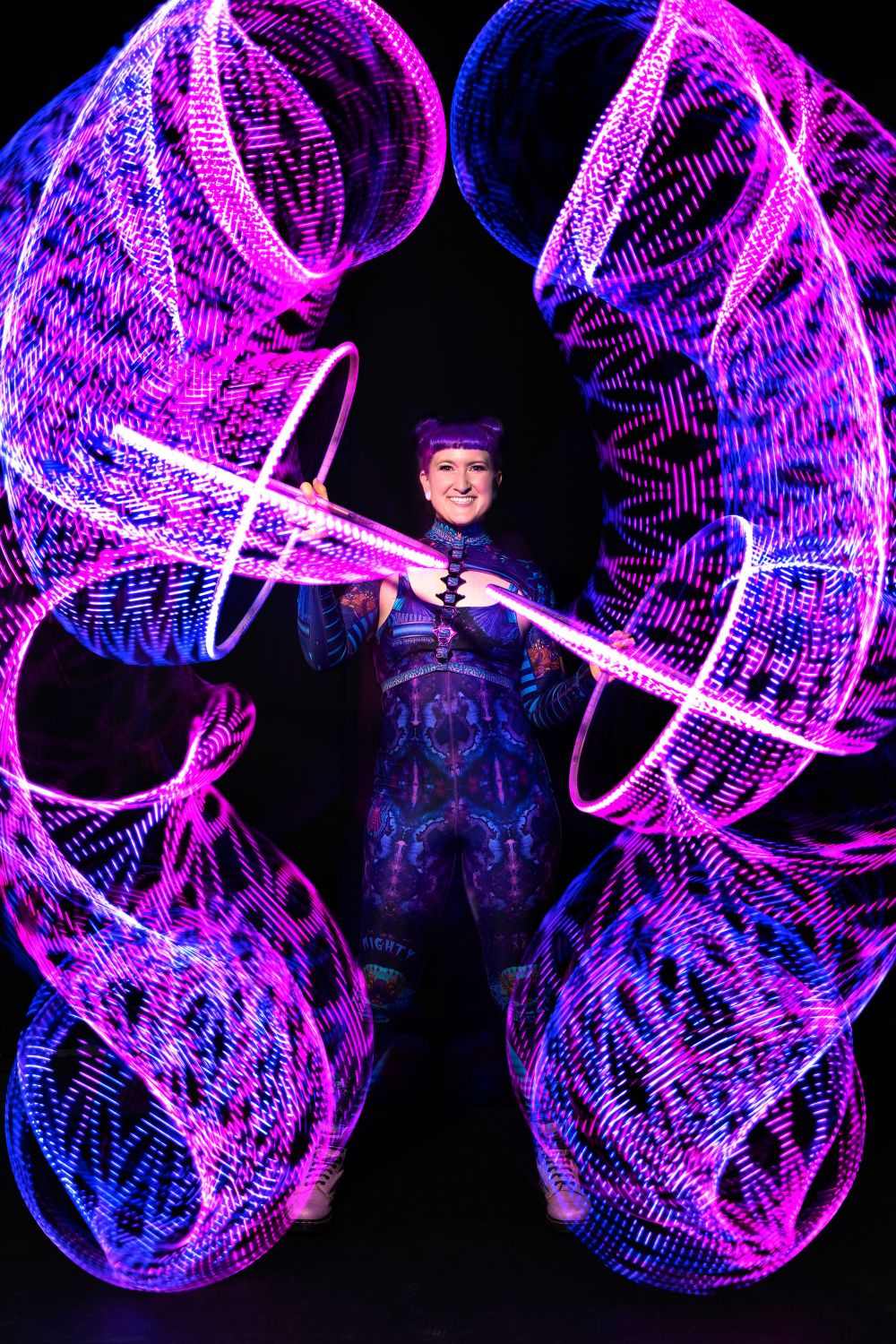 Long-exposure light painting: pink and blue hoop trails spiralling either side of Jess