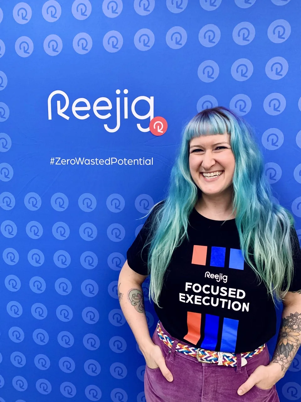 Jess smiling in front of a blue Reejig brand wall, wearing a Reejig Focused Execution t-shirt.
