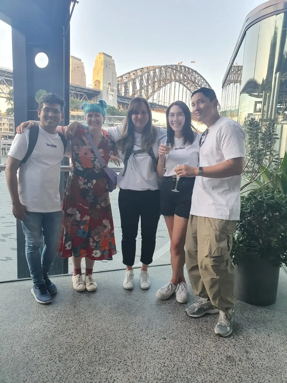 Five Reejig team members, including Jess, standing arm in arm on a waterfront terrace in Sydney with the Harbour Bridge behind them.